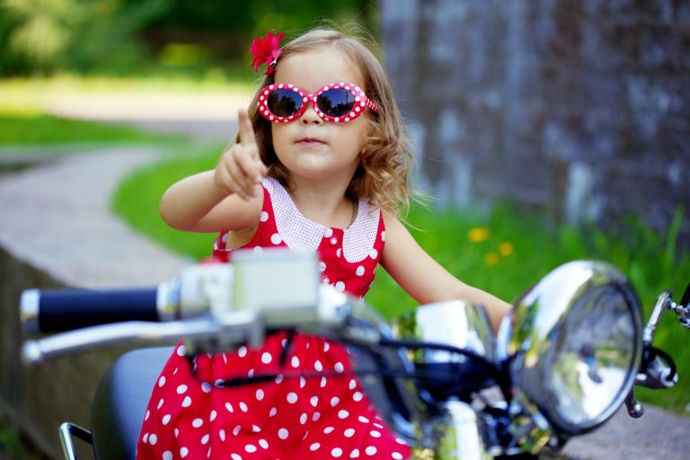 Child aged about 4 with red polkadot party dress and matching sunglasses, sitting on a motorbike.