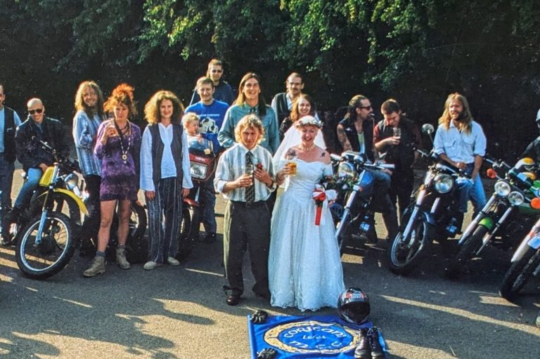 Male and female couple wearing wedding outfits with bikers and their motorbikes forming a circle around them.